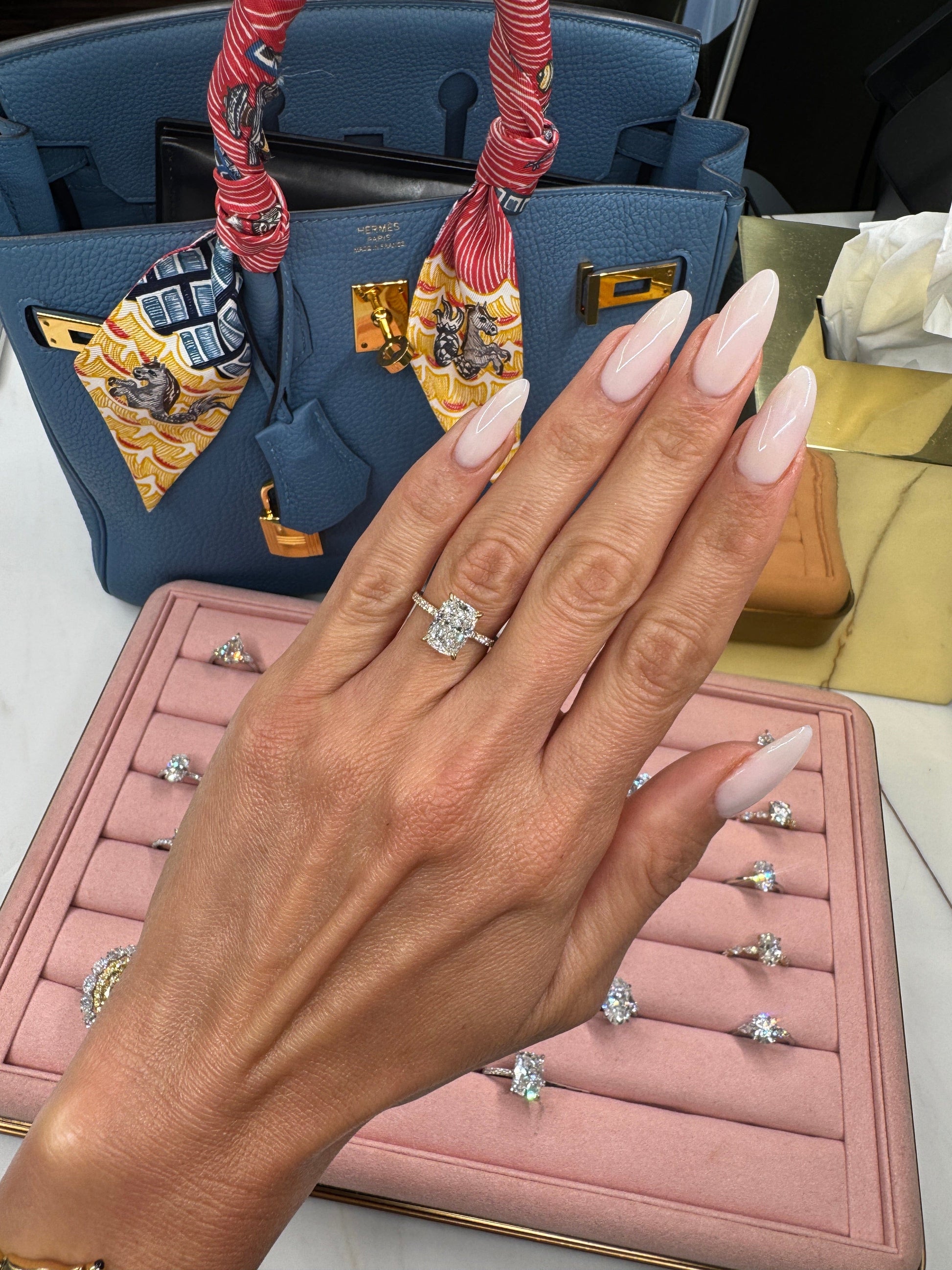 Hand with engagement ring, display of other rings, blue handbag.